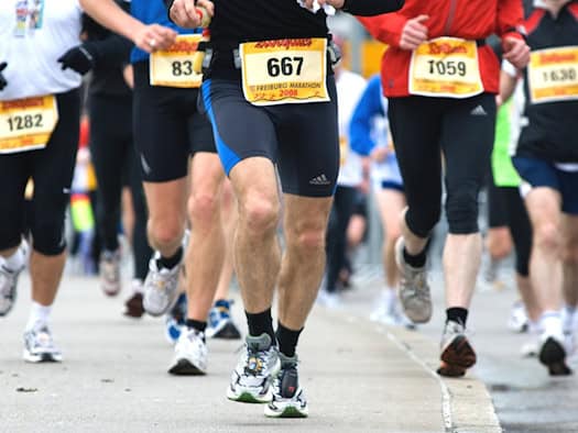 Nahaufnahme der Beine und Hüften mehrerer Läuferinnen und Läufer auf einer Asphaltstraße. Die Athleten tragen Sportkleidung, Laufschuhe und gelbe Startnummern an den Hosen.
