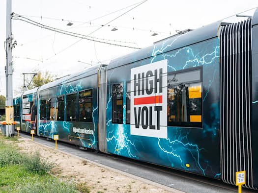 Perspektivische Außenaufnahme einer Straßenbahn auf den Gleisen. Die Bahn ist mit dunkler Folie, leuchtend blauen Blitzen, dem roten und weißen HIGHVOLT-Logo sowie dem Schriftzug „highvolt.com/karriere“ foliert.