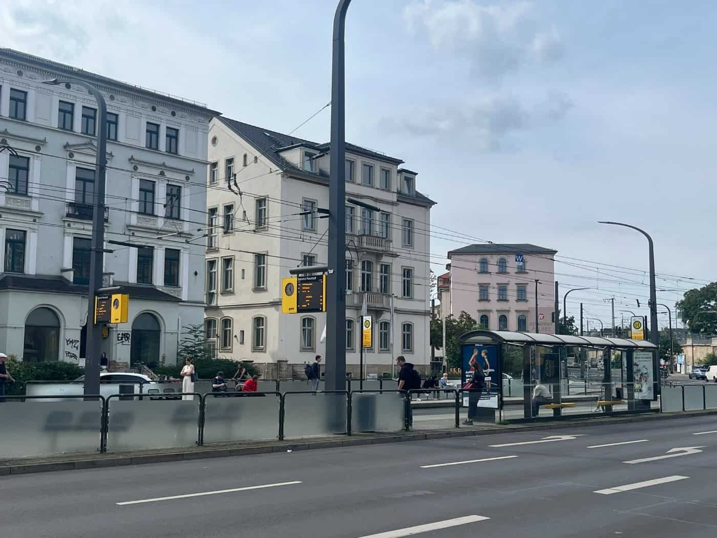 Eine mehrspurige Straße in Dresden mit der DVB-Straßenbahnhaltestelle Bahnhof Neustadt. Zu sehen sind Unterstand, gelbe Abfahrtsanzeigen und wartende Fahrgäste vor mehrstöckigen Gründerzeitgebäuden unter leicht bewölktem Himmel.