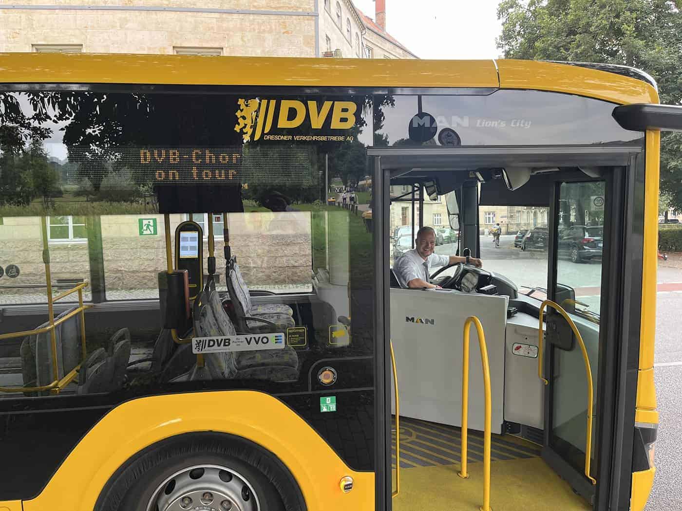 Seitenansicht eines gelben DVB-Busses mit der digitalen Anzeige DVB-Chor on tour. Durch die geöffnete Vordertür blickt der lächelnde Busfahrer am Steuer direkt in die Kamera.