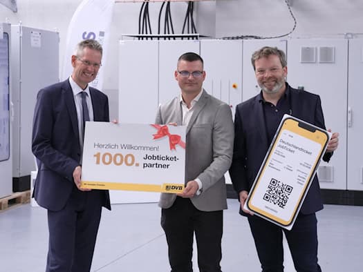 Drei Männer in Anzügen stehen lächelnd in einer Industriehalle vor grau-weißen Schaltschränken. Zwei halten ein Schild mit der Aufschrift „1000. Jobticket-partner“, der Rechte hält ein großes Schild in Form eines Smartphones mit digitalem Deutschlandticket.