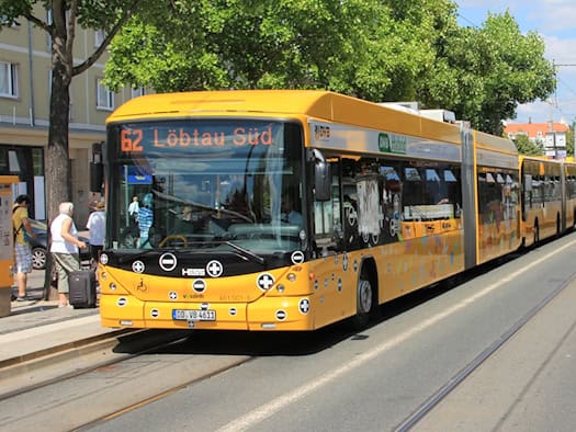 Ein gelber Doppelgelenkbus der DVB mit der Aufschrift „62 Löbtau Süd“ fährt an einer Haltestelle entlang. Daneben steht eine gelbe Straßenbahn auf Gleisen. Im Hintergrund befinden sich grüne Bäume und ein sommerlicher blauer Himmel mit leichten Wolken.