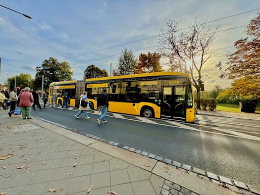 Gelb-schwarzer DVB-Gelenk-Elektrobus steht an einer Dresdner Straße in der Herbstsonne. Passanten überqueren die Straße vor dem Bus, auf dem die Aufschrift „Wir haben neue E-Busse!“ zu sehen ist.