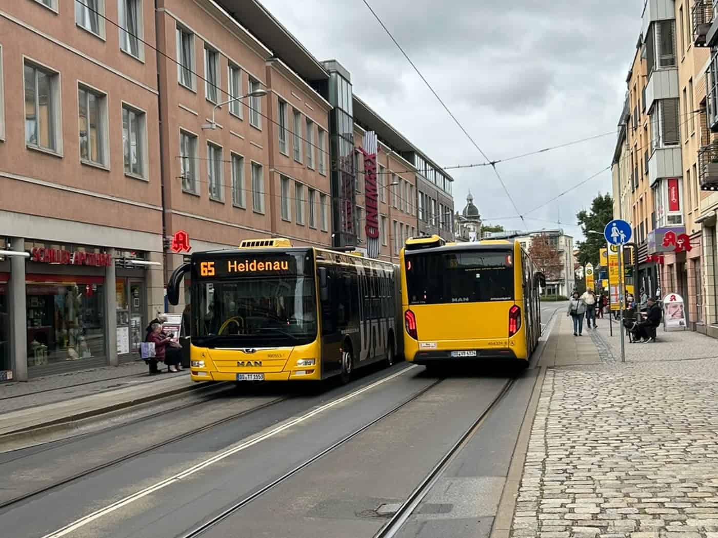 Ein gelber DVB-Bus der Linie 65 mit der Zielanzeige Heidenau fährt auf einer Straße mit Straßenbahnschienen an einem weiteren gelben Bus vorbei, gesäumt von mehrstöckigen Wohn- und Geschäftshäusern.