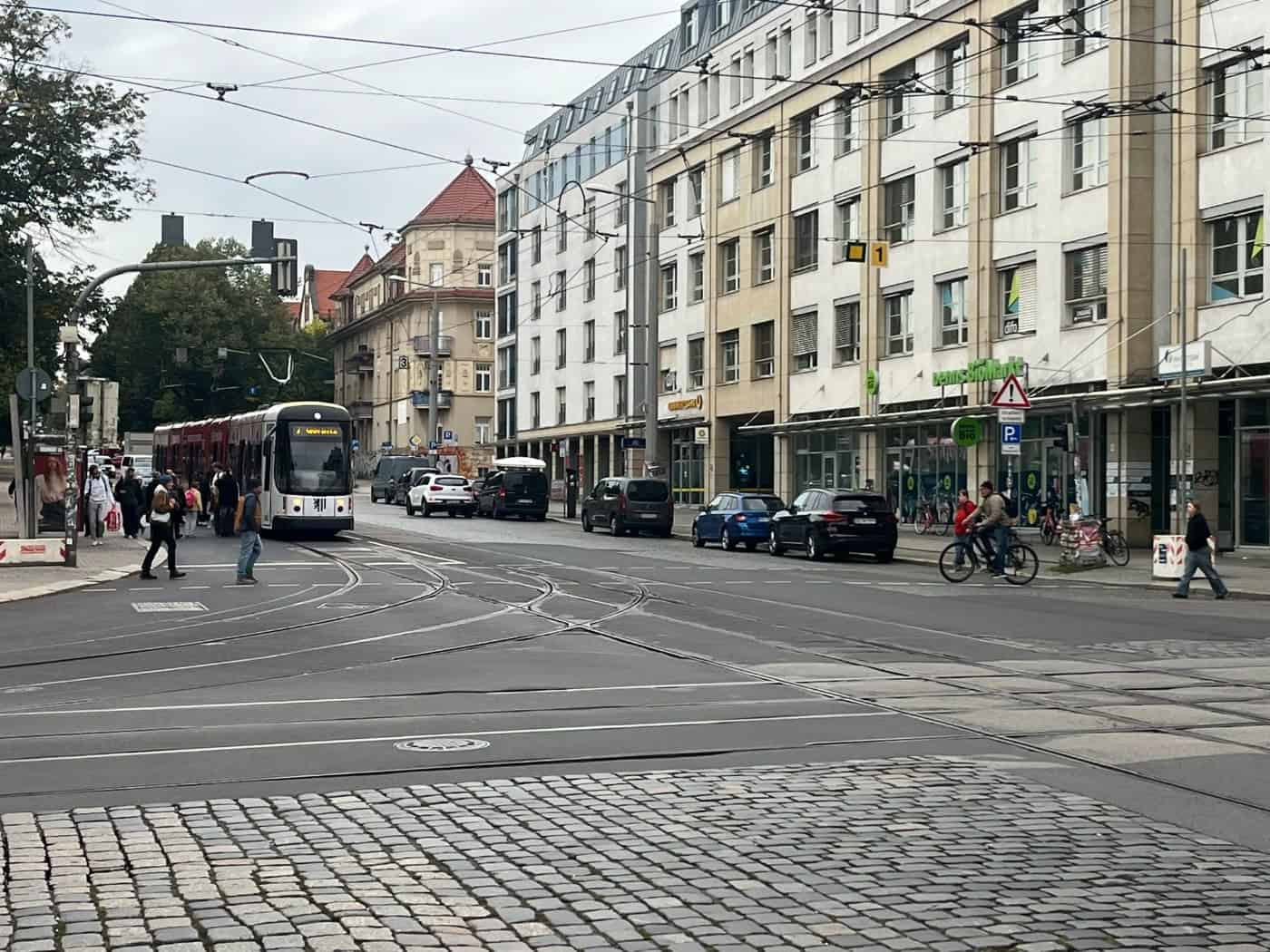 Eine gelb-schwarze DVB-Straßenbahn überquert die Kreuzung Königsbrücker Straße und Bischofsweg in der Dresdner Neustadt. Im Vordergrund verlaufen Straßenbahnschienen auf der Straße, im Hintergrund stehen mehrgeschossige Wohn- und Geschäftshäuser.