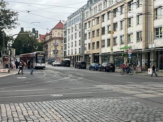 Eine gelb-schwarze DVB-Straßenbahn überquert die Kreuzung Königsbrücker Straße und Bischofsweg in der Dresdner Neustadt. Im Vordergrund verlaufen Straßenbahnschienen auf der Straße, im Hintergrund stehen mehrgeschossige Wohn- und Geschäftshäuser.