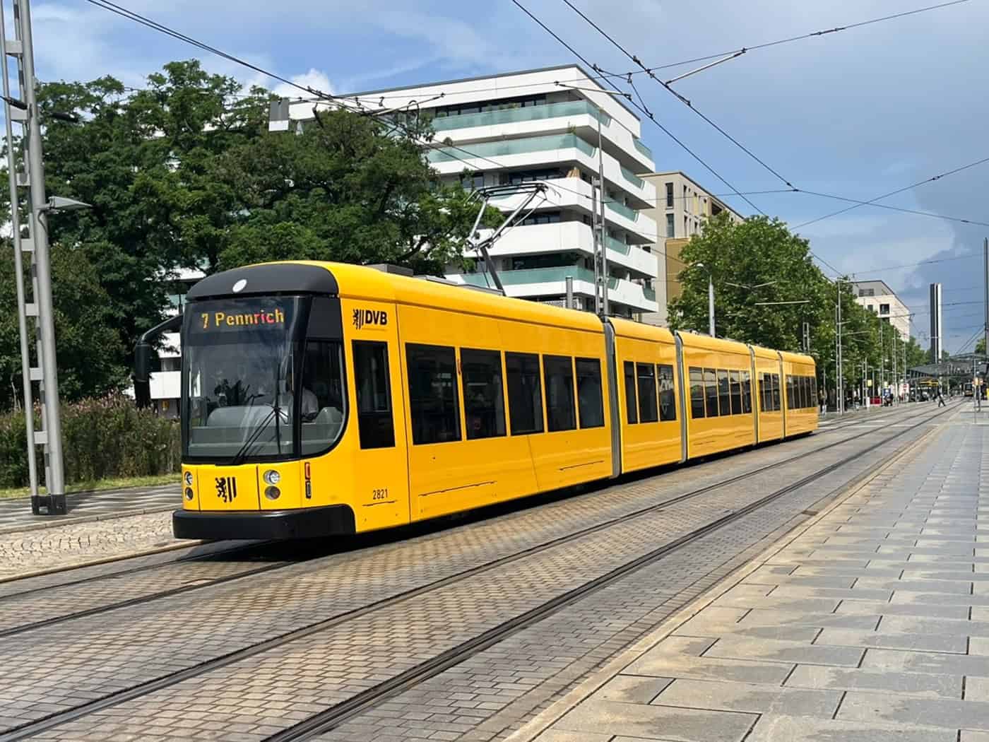 Eine gelbe Straßenbahn der Dresdner Verkehrsbetriebe (DVB) der Linie 7 mit der Zielanzeige Pennrich fährt auf Schienen über eine gepflasterte Straße, vorbei an moderneren Gebäuden unter leicht bewölktem Himmel.