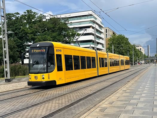 Eine gelbe Straßenbahn der Dresdner Verkehrsbetriebe (DVB) der Linie 7 mit der Zielanzeige Pennrich fährt auf Schienen über eine gepflasterte Straße, vorbei an moderneren Gebäuden unter leicht bewölktem Himmel.