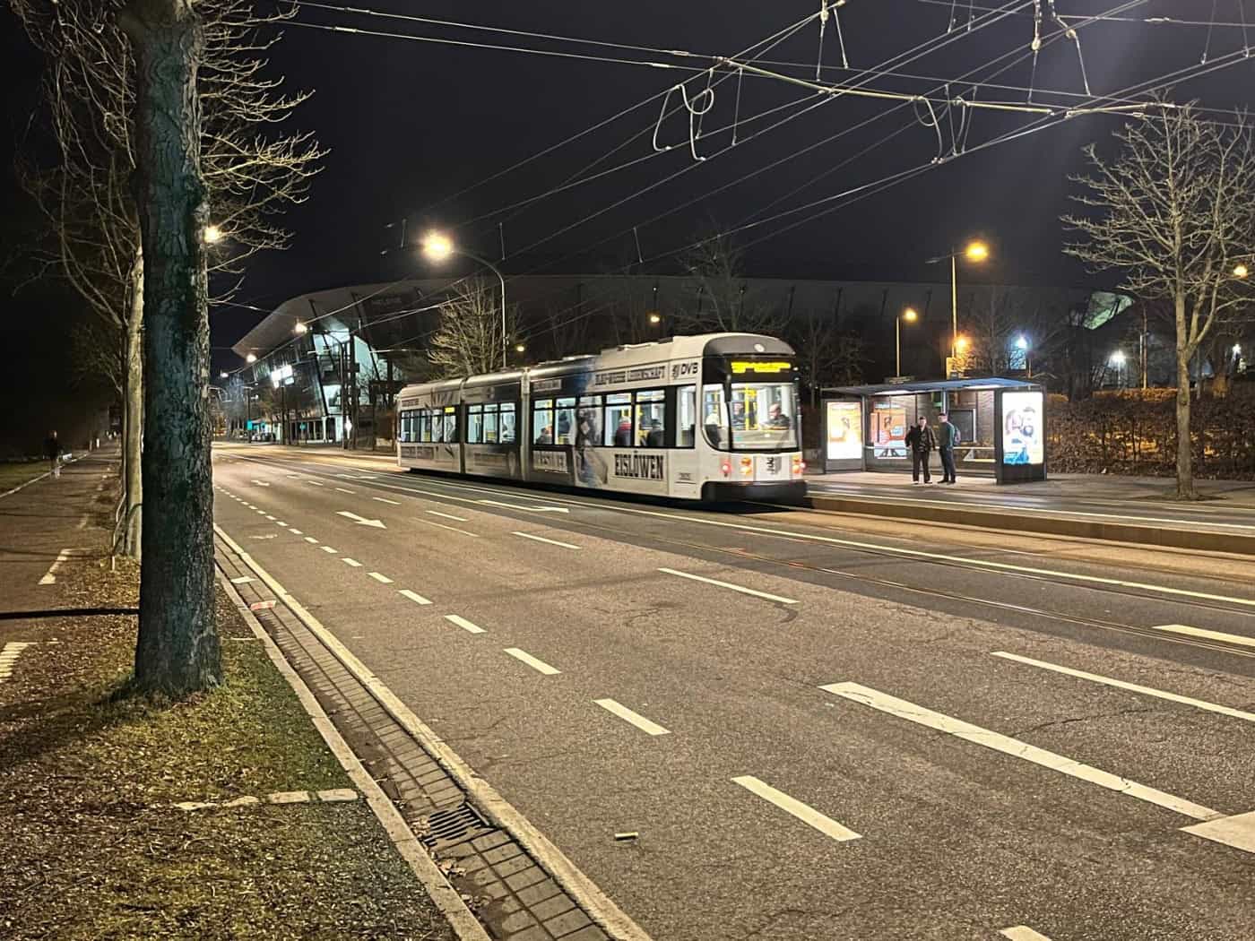 Nächtliche Straßenansicht mit einer gelb-weißen DVB-Straßenbahn an einer Haltestelle. Im dunklen Hintergrund ist die beleuchtete Silhouette des Rudolf-Harbig-Stadions in Dresden erkennbar. Zwei Personen warten im Unterstand.