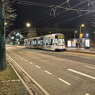 Nächtliche Straßenansicht mit einer gelb-weißen DVB-Straßenbahn an einer Haltestelle. Im dunklen Hintergrund ist die beleuchtete Silhouette des Rudolf-Harbig-Stadions in Dresden erkennbar. Zwei Personen warten im Unterstand.