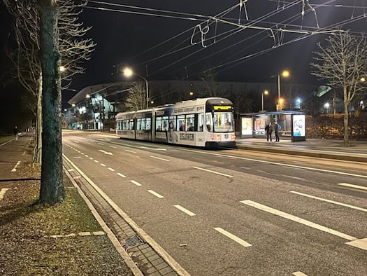 Nächtliche Straßenansicht mit einer gelb-weißen DVB-Straßenbahn an einer Haltestelle. Im dunklen Hintergrund ist die beleuchtete Silhouette des Rudolf-Harbig-Stadions in Dresden erkennbar. Zwei Personen warten im Unterstand.