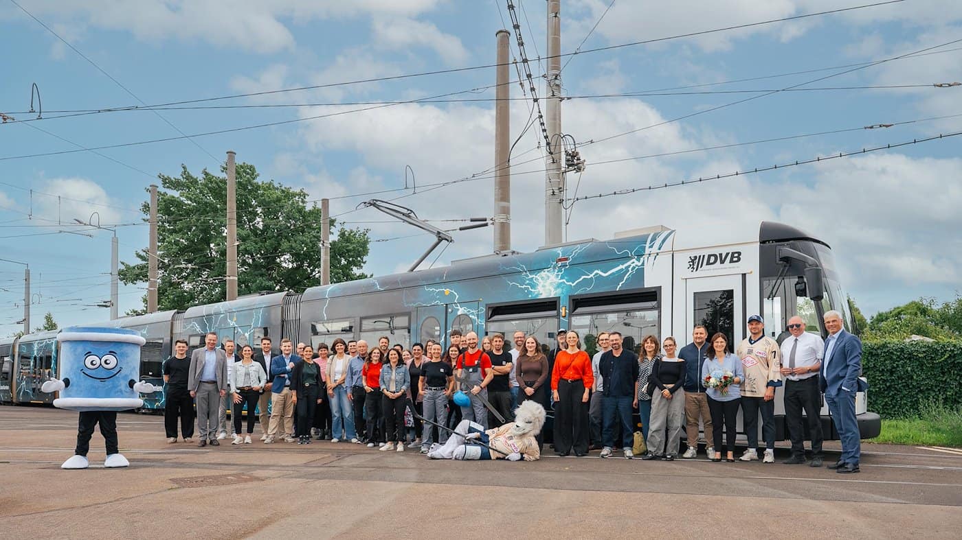 Große Personengruppe und zwei Maskottchen stehen lächelnd vor einer Straßenbahn der Dresdner Verkehrsbetriebe (DVB). Die Bahn ist im HIGHVOLT-Design mit blauen Blitzen bedruckt. Das Foto entstand auf einem Betriebshof unter leicht bewölktem Himmel.