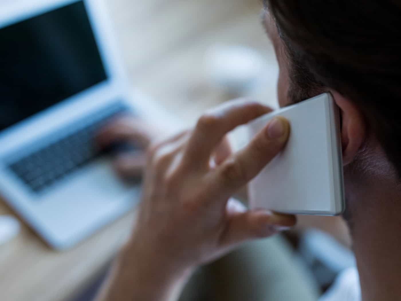 Nahaufnahme von hinten: Ein Mann hält ein weißes Smartphone an sein Ohr und bedient gleichzeitig mit der anderen Hand die Tastatur eines unscharf im Hintergrund stehenden Laptops.
