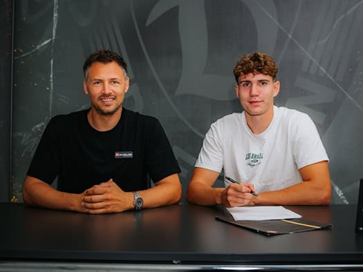 Sportgeschäftsführer Sören Gonther im schwarzen T-Shirt und Profi Friedrich Müller im weißen T-Shirt sitzen lächelnd nebeneinander an einem dunklen Tisch. Müller hält einen Stift über den Vertrag. Im Hintergrund befindet sich ein Dynamo-Dresden-Logo.