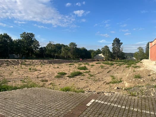 Weite, brachliegende Schotterfläche des ehemaligen Operettenareals in Dresden-Leuben. Im Vordergrund gepflasterter Parkplatzboden, im Hintergrund eine Baumreihe unter leicht bewölktem blauem Himmel und rechts eine rote Gebäudewand.