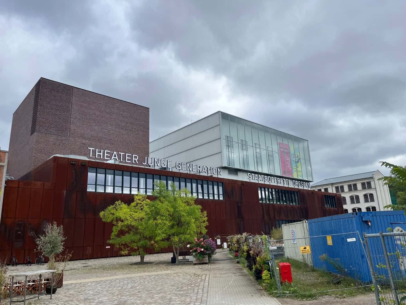 Außenansicht des modernen Gebäudes im Kraftwerk Mitte mit rostroter Fassade. Groß gedruckt stehen die Schriftzüge „THEATER JUNGE GENERATION“ und „STAATSOPERETTE DRESDEN“ an der Front. Davor ein Vorplatz mit Bäumen unter bedecktem Himmel.