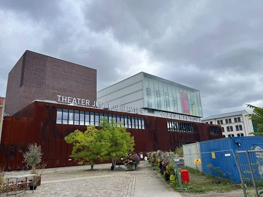 Außenansicht des modernen Gebäudes im Kraftwerk Mitte mit rostroter Fassade. Groß gedruckt stehen die Schriftzüge „THEATER JUNGE GENERATION“ und „STAATSOPERETTE DRESDEN“ an der Front. Davor ein Vorplatz mit Bäumen unter bedecktem Himmel.