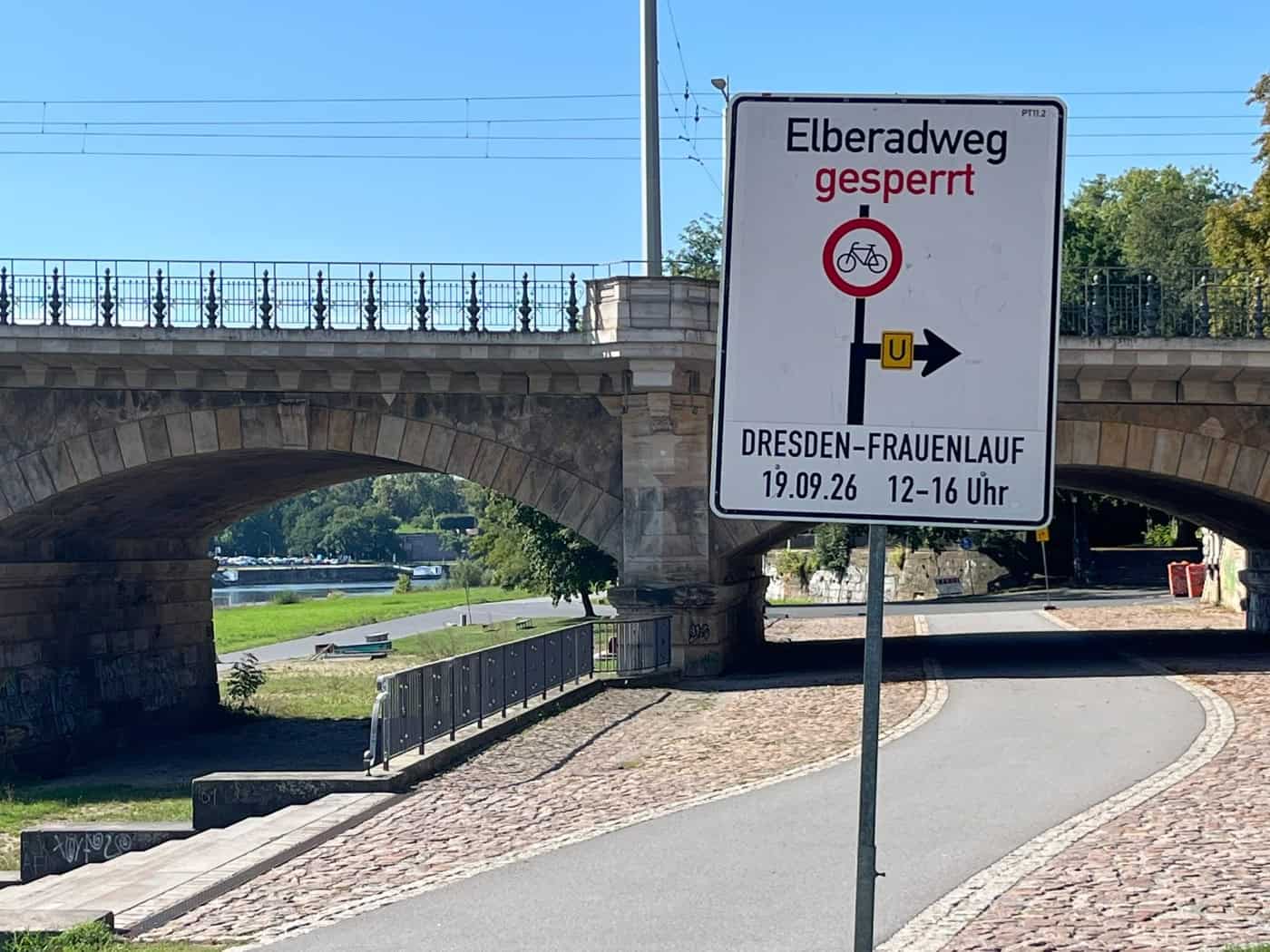 Ein weißes Hinweisschild mit der Aufschrift „Elberadweg gesperrt“, einem Fahrradverbotssymbol und dem Hinweis „DRESDEN-FRAUENLAUF 19.09.26 12–16 Uhr“ steht vor einer Sandsteinbrücke am Elberadweg.