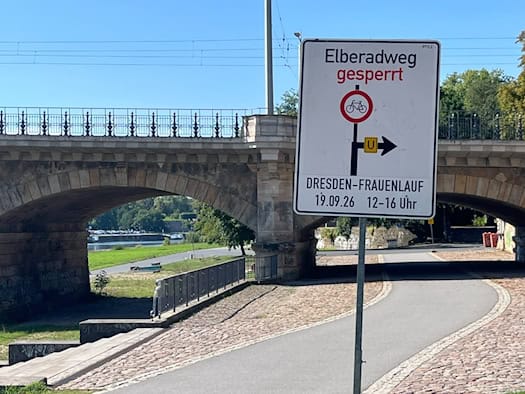 Ein weißes Hinweisschild mit der Aufschrift „Elberadweg gesperrt“, einem Fahrradverbotssymbol und dem Hinweis „DRESDEN-FRAUENLAUF 19.09.26 12–16 Uhr“ steht vor einer Sandsteinbrücke am Elberadweg.