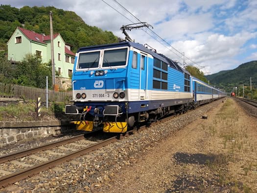 Ein blau-weißer Eurocity-Zug der Tschechischen Bahnen mit Elektrolok fährt auf Gleisen durch eine grüne Landschaft an Wohnhäusern vorbei. Symbolbild für die Strecke Prag–Dresden, auf der die Bundespolizei bei Kontrollen einen Gesuchten stellte.