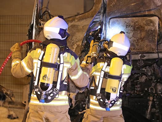 Zwei Feuerwehrleute in Schutzkleidung mit gelben Atemschutzflaschen auf dem Rücken arbeiten in der Nacht an der verkohlten Fahrerkabine eines Lkw. Einer hält einen roten Schlauch, während sie die Brandstelle am Fahrzeug sichern.