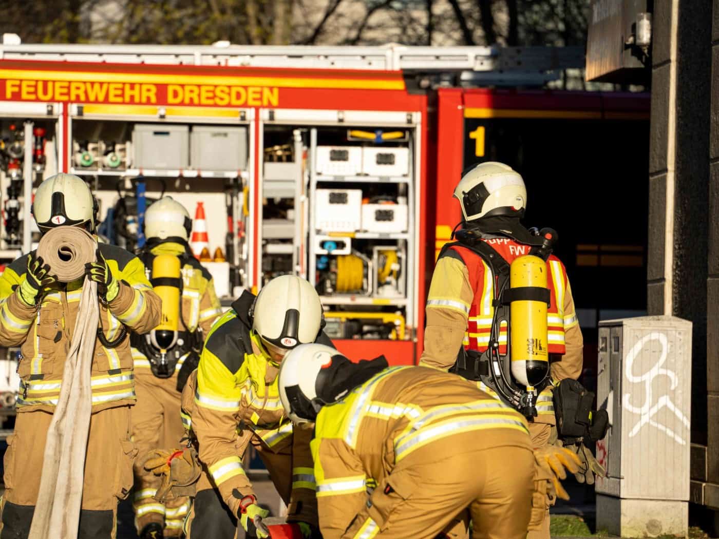 Mehrere Einsatzkräfte der Feuerwehr Dresden in Schutzkleidung und mit Atemschutzgeräten vor einem roten Einsatzfahrzeug. Ein Feuerwehrmann im Vordergrund hält einen gerollten Löschschlauch, während sich andere Einsatzkräfte zu weiterem Material beugen.
