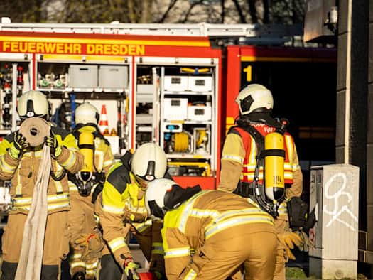 Mehrere Einsatzkräfte der Feuerwehr Dresden in Schutzkleidung und mit Atemschutzgeräten vor einem roten Einsatzfahrzeug. Ein Feuerwehrmann im Vordergrund hält einen gerollten Löschschlauch, während sich andere Einsatzkräfte zu weiterem Material beugen.