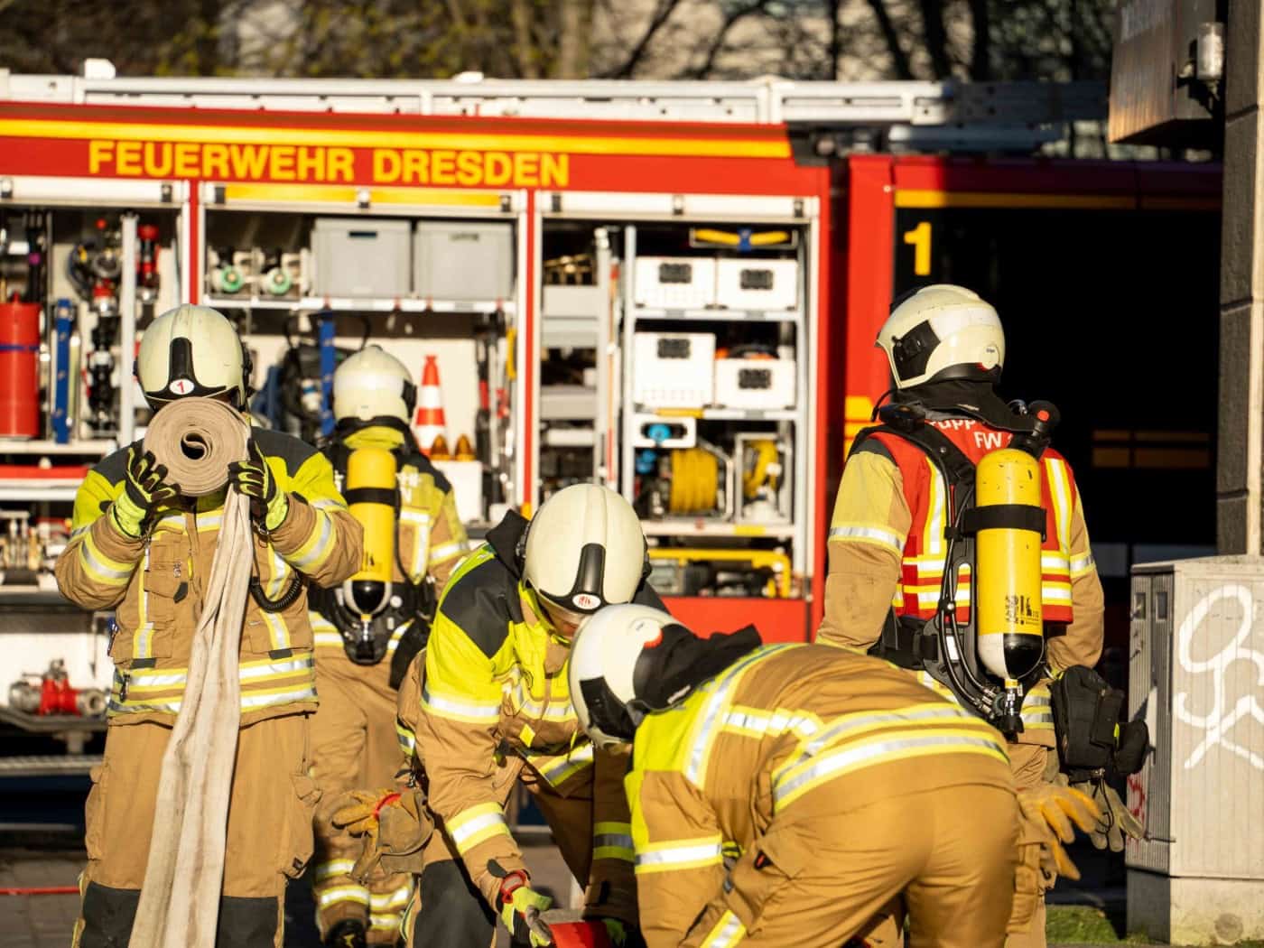 Mehrere Feuerwehrleute in schützender Einsatzkleidung mit gelben Atemschutzgeräten bereiten vor einem roten Einsatzfahrzeug der Feuerwehr Dresden Schläuche vor. Im Hintergrund ist ein geparktes Löschfahrzeug mit geöffneten Geräteräumen zu sehen.