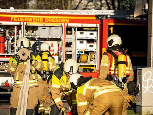 Mehrere Feuerwehrleute in schützender Einsatzkleidung mit gelben Atemschutzgeräten bereiten vor einem roten Einsatzfahrzeug der Feuerwehr Dresden Schläuche vor. Im Hintergrund ist ein geparktes Löschfahrzeug mit geöffneten Geräteräumen zu sehen.