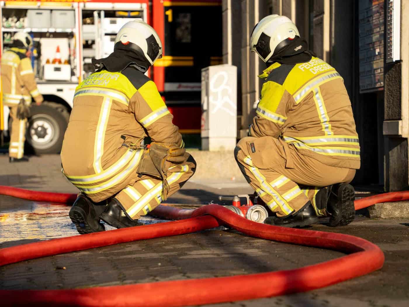 Symbolbild: Zwei Feuerwehrleute in beige-gelber Schutzkleidung hocken auf dem Gehweg an roten Feuerwehrschläuchen. Auf der Jacke steht „FEUERWEHR DRESDEN“. Im unscharfen Hintergrund ist ein rotes Feuerwehrfahrzeug zu sehen.