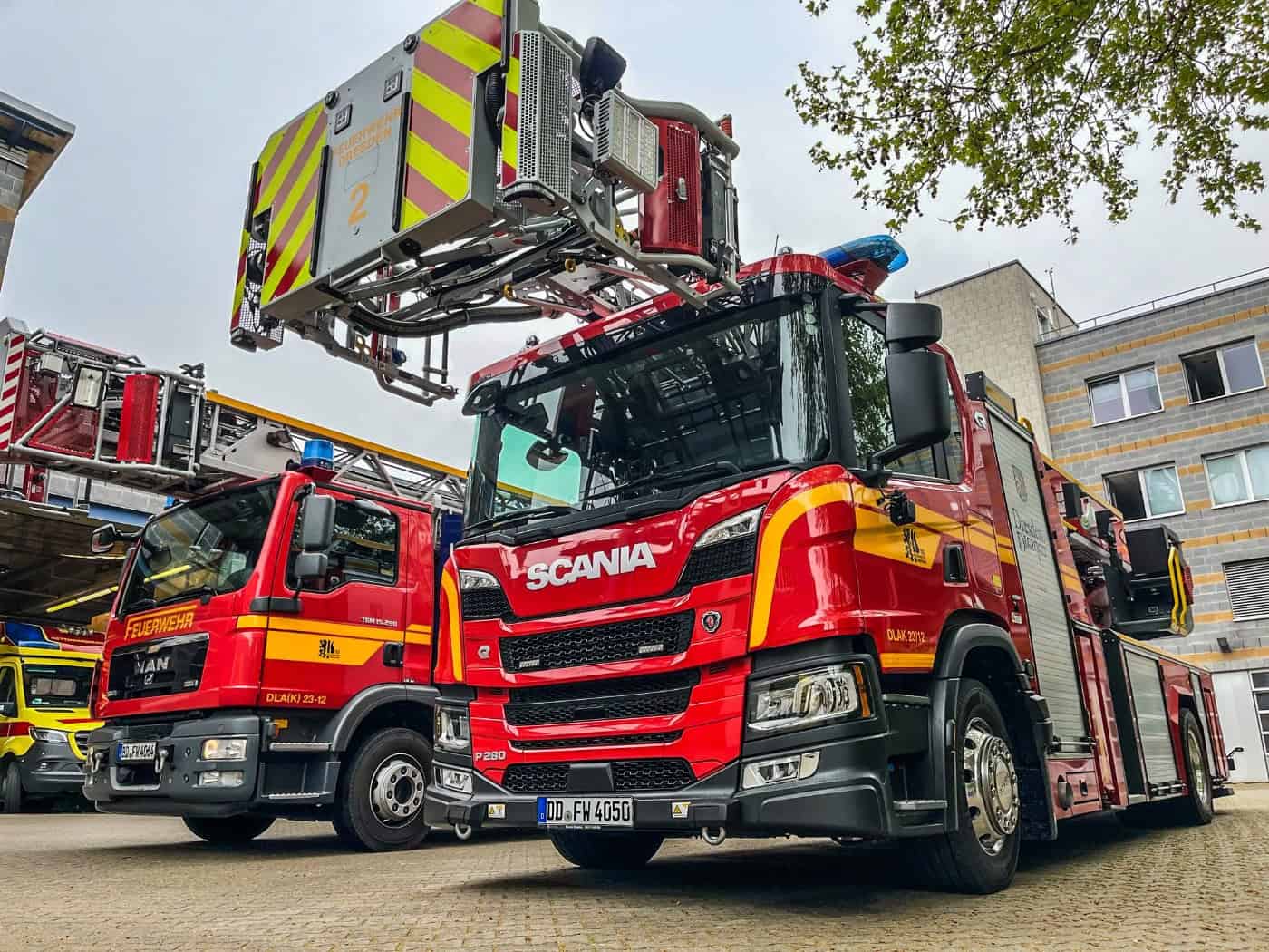 Rot-gelbes Drehleiterfahrzeug der Marke Scania von der Feuerwehr Dresden steht auf einem Betriebshof. Daneben parkt ein weiteres rotes Feuerwehrfahrzeug, ganz links ist der Teil eines gelben Rettungswagens zu sehen.