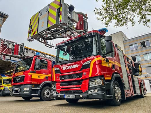 Rot-gelbes Drehleiterfahrzeug der Marke Scania von der Feuerwehr Dresden steht auf einem Betriebshof. Daneben parkt ein weiteres rotes Feuerwehrfahrzeug, ganz links ist der Teil eines gelben Rettungswagens zu sehen.