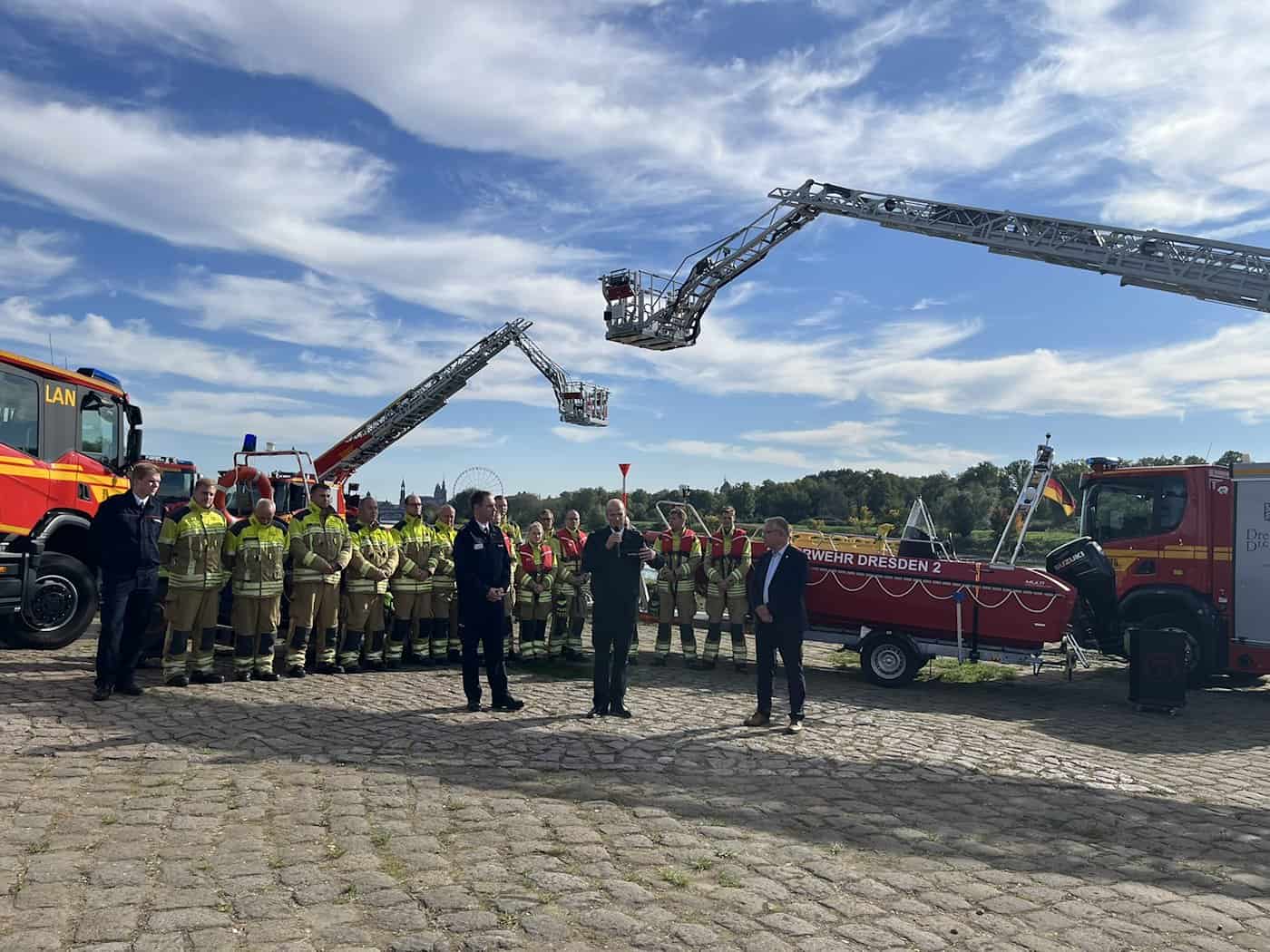 Panorama am Dresdner Elbufer: Feierliche Übergabe neuer Einsatzfahrzeuge mit ausgefahrenen Drehleitern, Löschfahrzeugen und Rettungsbooten vor aufgestellten Feuerwehrleuten.