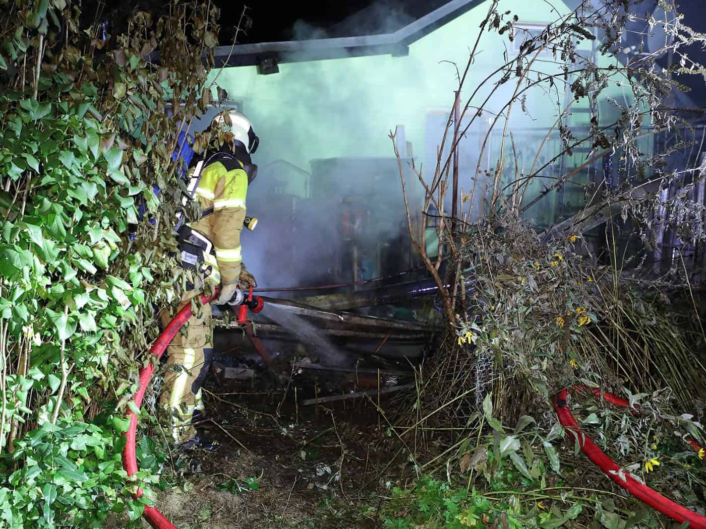 Nächtlicher Löscheinsatz: Ein Feuerwehrmann in Schutzkleidung und Atemschutz richtet einen Wasserstrahl aus einem roten Schlauch auf rauchende, verbrannte Holztrümmer zwischen dichtem Gebüsch in einer Kleingartenanlage.   