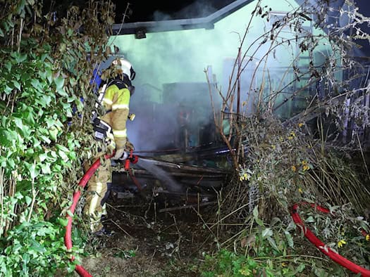 Nächtlicher Löscheinsatz: Ein Feuerwehrmann in Schutzkleidung und Atemschutz richtet einen Wasserstrahl aus einem roten Schlauch auf rauchende, verbrannte Holztrümmer zwischen dichtem Gebüsch in einer Kleingartenanlage.   