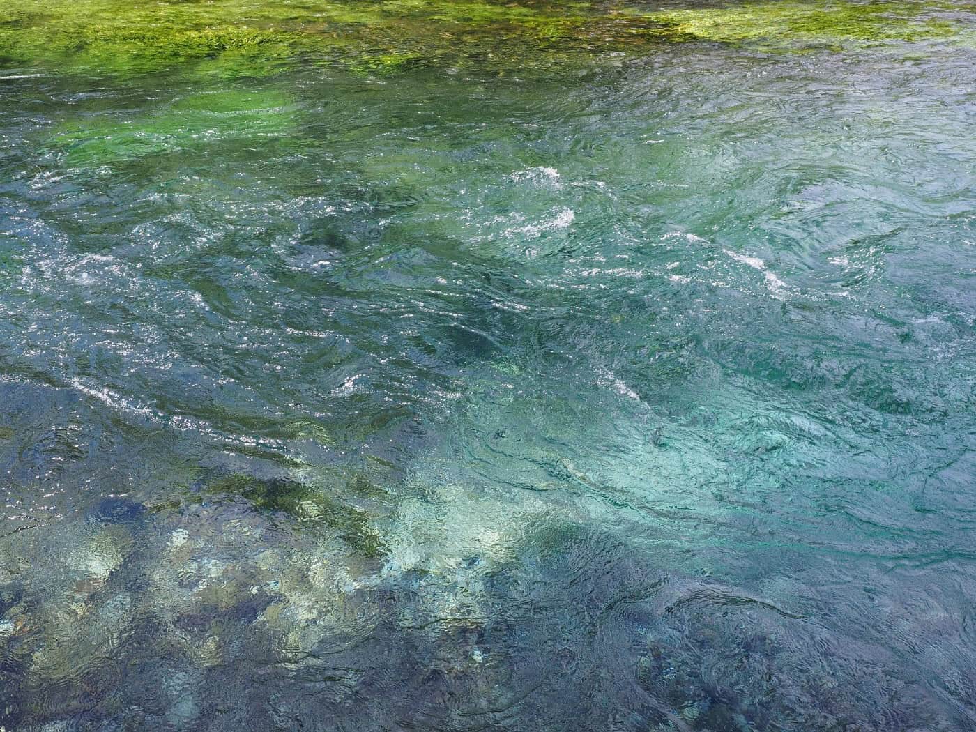 Nahaufnahme von klarem, grünlich-blau schimmerndem Wasser eines Baches, das mit leichten Wellen und Strömungswirbeln fließt. Am Rand und auf dem Grund sind Kieselsteine und grünliche Wasserpflanzen unter der Wasseroberfläche zu erkennen.