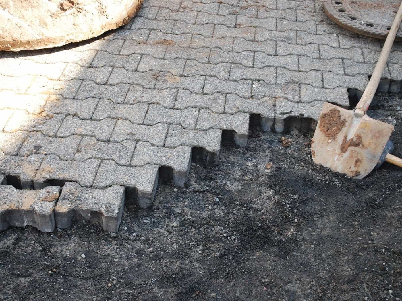 Nahaufnahme einer Baustelle: Ein Weg wird mit grauen Betonpflastersteinen verlegt. Neben den bereits gesetzten Steinen liegen Erde und ein dunkler Untergrund, auf dem eine verschmutzte Schaufel mit Holzstiel ruht. Rechts oben ist ein Kanaldeckel zu sehen.
