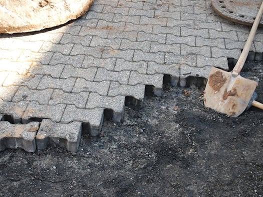 Nahaufnahme einer Baustelle: Ein Weg wird mit grauen Betonpflastersteinen verlegt. Neben den bereits gesetzten Steinen liegen Erde und ein dunkler Untergrund, auf dem eine verschmutzte Schaufel mit Holzstiel ruht. Rechts oben ist ein Kanaldeckel zu sehen.