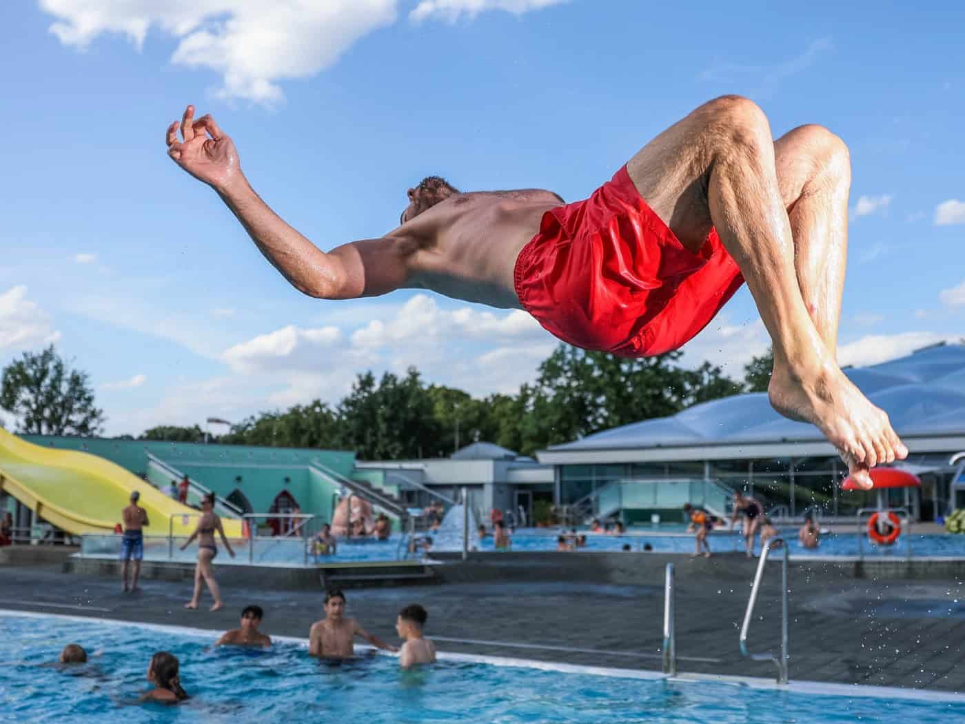 Ein Mann in roter Badehose macht einen Rückwärtssprung über dem blauen Wasser eines Freibads. Im Hintergrund sind Badegäste, eine gelbe Wasserrutsche und das Gebäude des Georg-Arnhold-Bads unter blauem Sommerhimmel zu sehen.