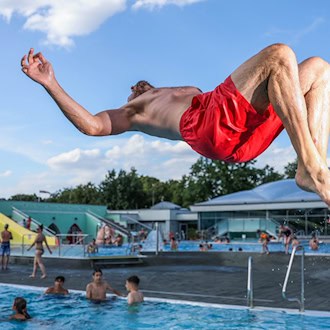 Ein Mann in roter Badehose macht einen Rückwärtssprung über dem blauen Wasser eines Freibads. Im Hintergrund sind Badegäste, eine gelbe Wasserrutsche und das Gebäude des Georg-Arnhold-Bads unter blauem Sommerhimmel zu sehen.
