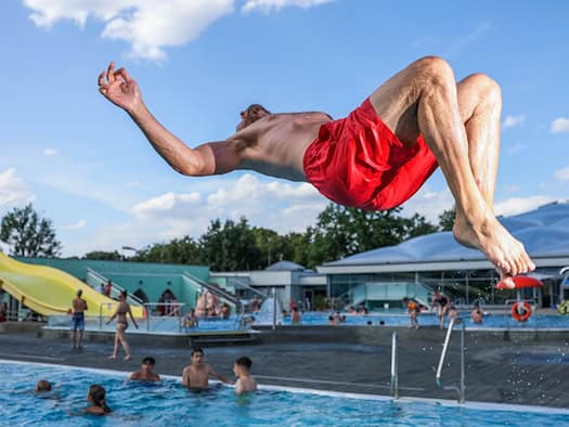 Ein Mann in roter Badehose macht einen Rückwärtssprung über dem blauen Wasser eines Freibads. Im Hintergrund sind Badegäste, eine gelbe Wasserrutsche und das Gebäude des Georg-Arnhold-Bads unter blauem Sommerhimmel zu sehen.