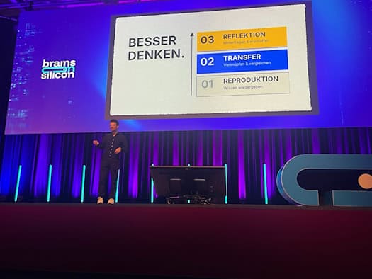 Gregor Schmalzried stands on the blue-lit stage at the “Brains on Silicon” conference. A screen in the background displays the “THINK BETTER” framework, with its three stages: reproduction, transfer, and reflection.