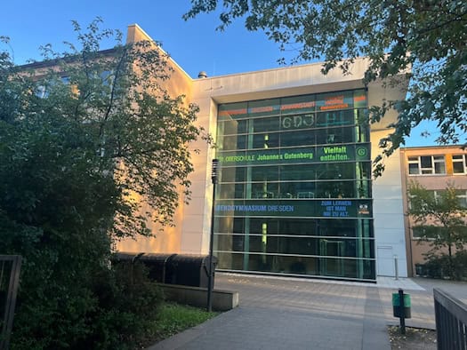 Moderne Glasfassade des Schulgebäudes vom Gymnasium Dresden-Johannstadt im Sonnenschein. Am Eingang sind Schriftzüge des Gymnasiums, der 101. Oberschule und des Abendgymnasiums zu sehen. Grüner Baumbestand umrahmt das Gebäude.