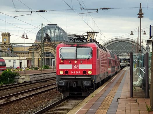 Eine rote Deutsche Bahn Lokomotive mit Doppelstockwagen steht an einem Bahnsteig des Dresdner Hauptbahnhofs. Im Hintergrund ist die große gläserne Kuppelhalle des historischen Bahnhofsgebäudes unter bewölktem Himmel zu sehen.