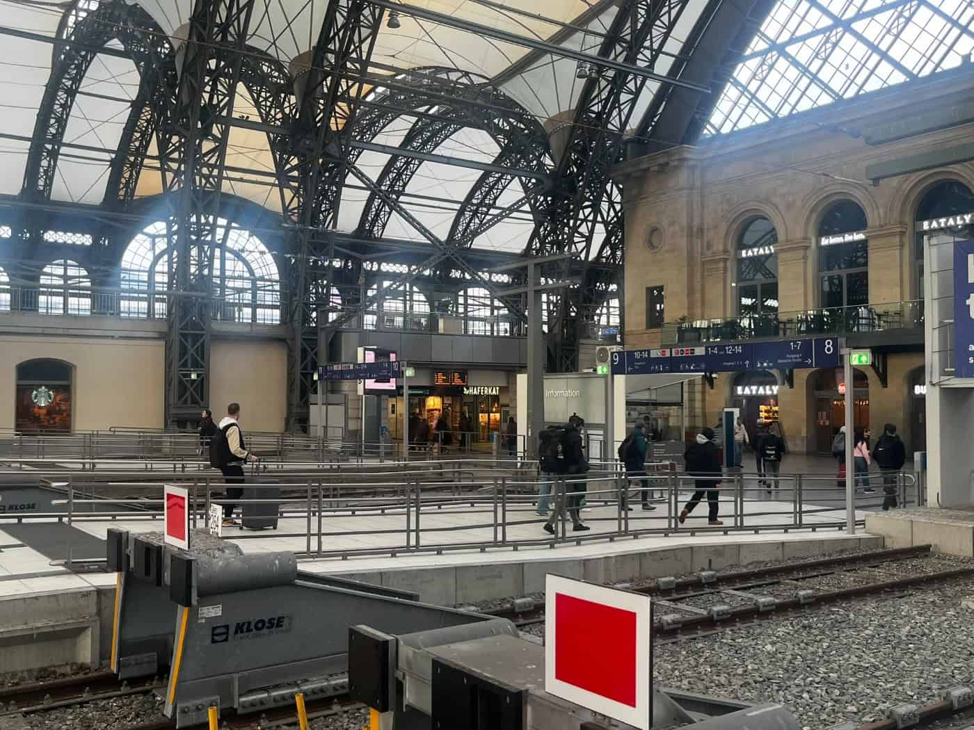 Innenansicht des Dresdner Hauptbahnhofs mit Gleisenden, Prellböcken, Fahrgästen auf dem Bahnsteig und Geschäften unter der imposanten Stahl- und Glaskuppel der Bahnhofshalle.