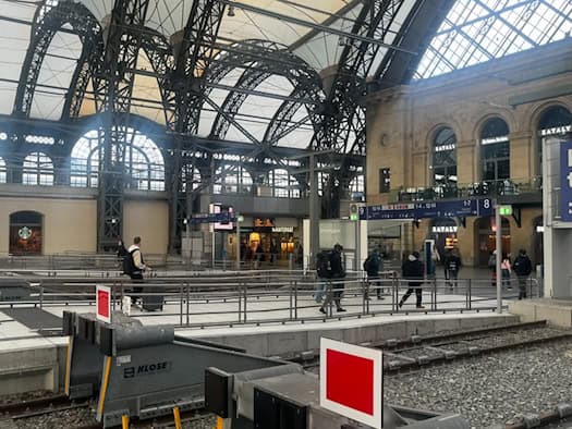 Innenansicht des Dresdner Hauptbahnhofs mit Gleisenden, Prellböcken, Fahrgästen auf dem Bahnsteig und Geschäften unter der imposanten Stahl- und Glaskuppel der Bahnhofshalle.