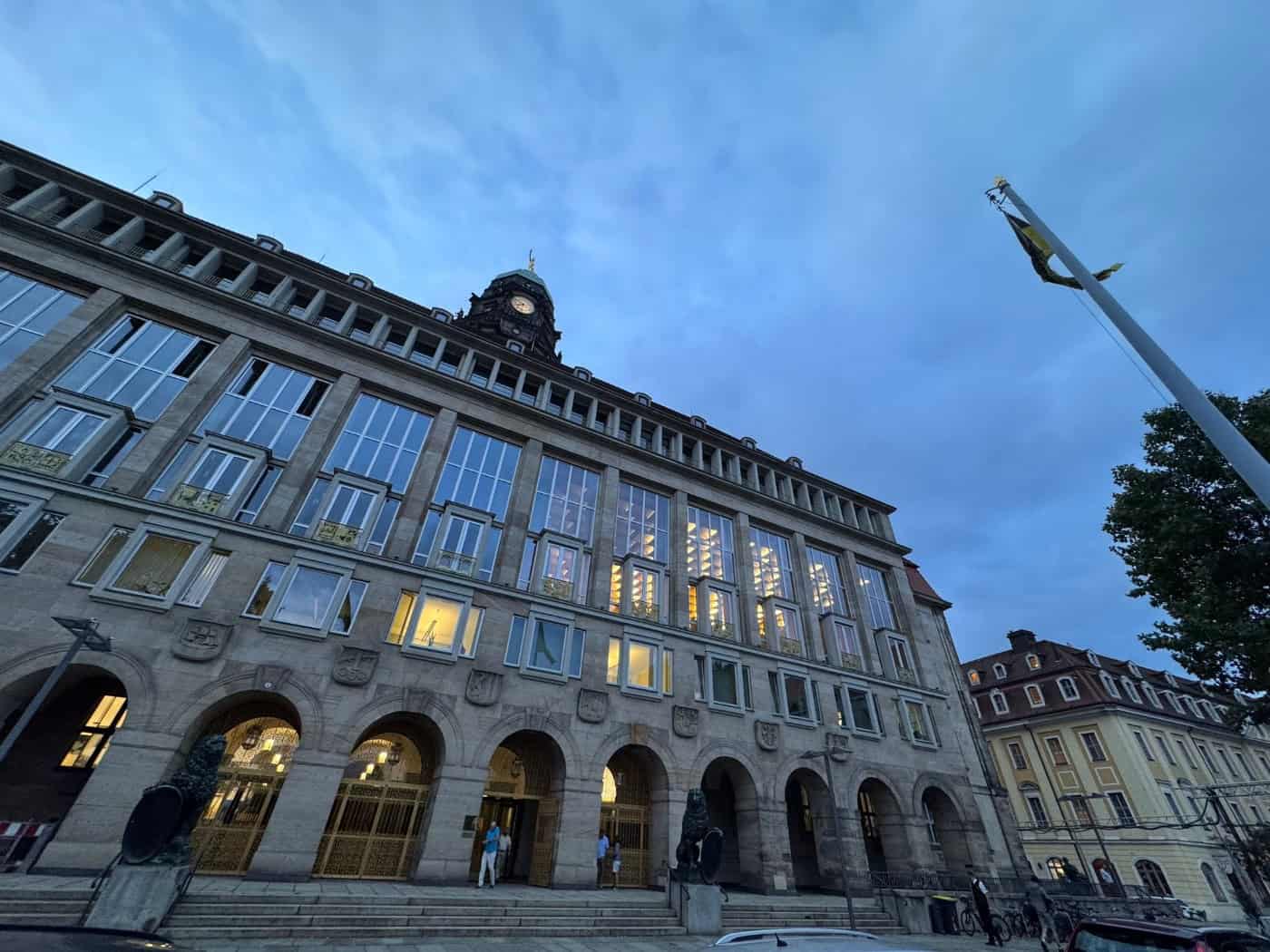 Froschperspektive auf die Fassade des Neuen Rathauses in Dresden bei Dämmerung. Die erleuchteten Rundbogen-Eingänge und Fenster heben sich vom dämmrigen blauen Abendhimmel ab. Rechts ragt ein Fahnenmast ins Bild.