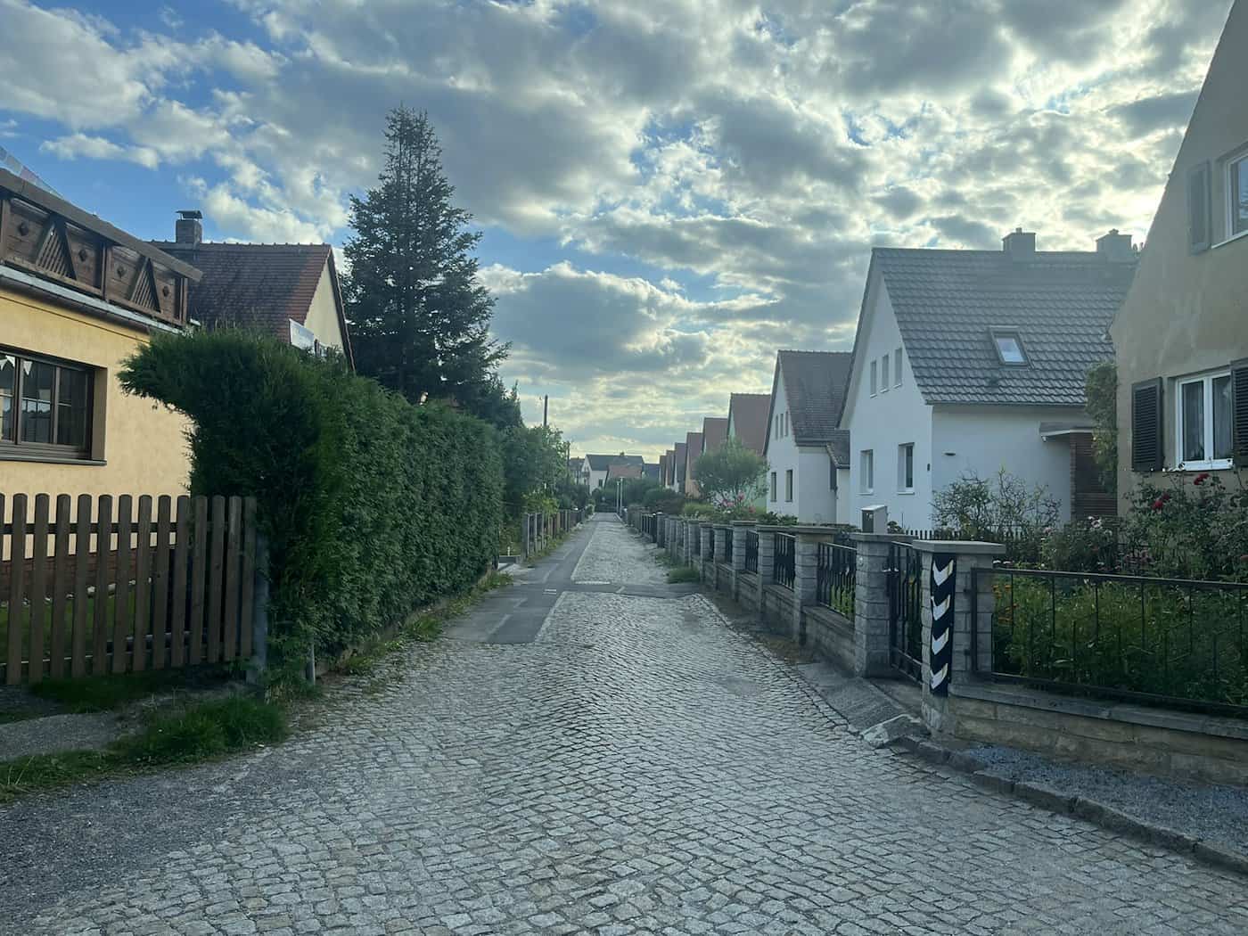 Blick durch eine schmale, mit Kopfsteinpflaster gepflasterte Wohnstraße in Dresden-Kleinzschachwitz. Zu beiden Seiten stehen Einfamilienhäuser mit Vorgärten und Zäunen unter einem leicht bewölkten blauen Himmel.
