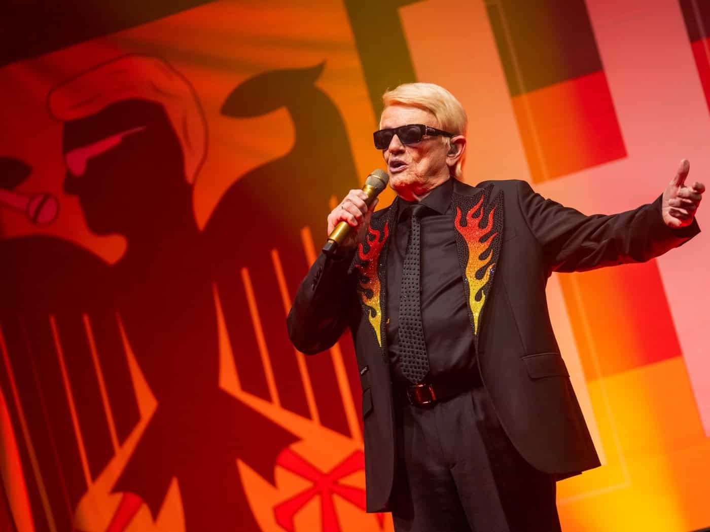 Sänger Heino steht mit blonder Frisur, dunkler Sonnenbrille und Flammenmuster auf dem Sakkorevers mit einem goldenen Mikrofon auf der Bühne. Im Hintergrund ist eine stilisierte Deutschlandflagge mit der Silhouette des Bundesadlers zu sehen.