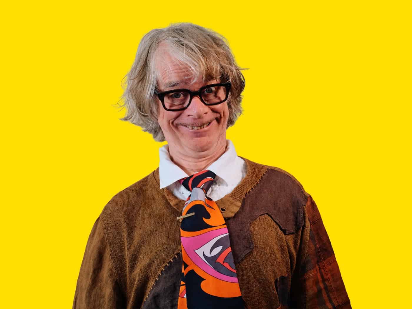 Porträt von Komiker Helge Schneider vor knallgelbem Hintergrund. Er schaut mit schwarzer Brille, grauer Perücke und schrägem Grinsen in die Kamera, trägt eine braune Strickjacke und eine bunt gemusterte Krawatte im 70er-Jahre-Stil.